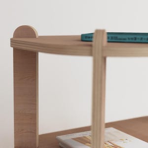 Puede incluir: Una mesa auxiliar de madera clara con dos estantes. Un libro turquesa y una peque&ntilde;a planta en maceta con hojas verdes est&aacute;n en el estante superior. La mesa tiene un dise&ntilde;o simple y moderno.