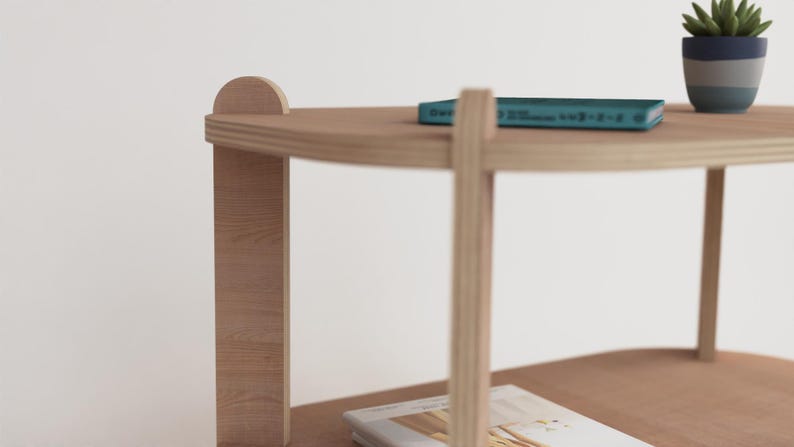 Puede incluir: Una mesa auxiliar de madera clara con dos estantes. Un libro turquesa y una peque&ntilde;a planta en maceta con hojas verdes est&aacute;n en el estante superior. La mesa tiene un dise&ntilde;o simple y moderno.