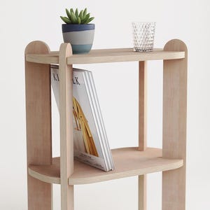 May include: A light-colored wooden shelf unit with three tiers. A small potted plant and a clear glass are on the top shelf. A magazine is placed on the middle shelf. The unit has a minimalist design.