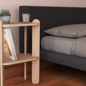 May include: A light wood bedside table with two shelves, holding a potted succulent, a glass, and a book. The table is next to a bed with gray bedding and a dark gray headboard.