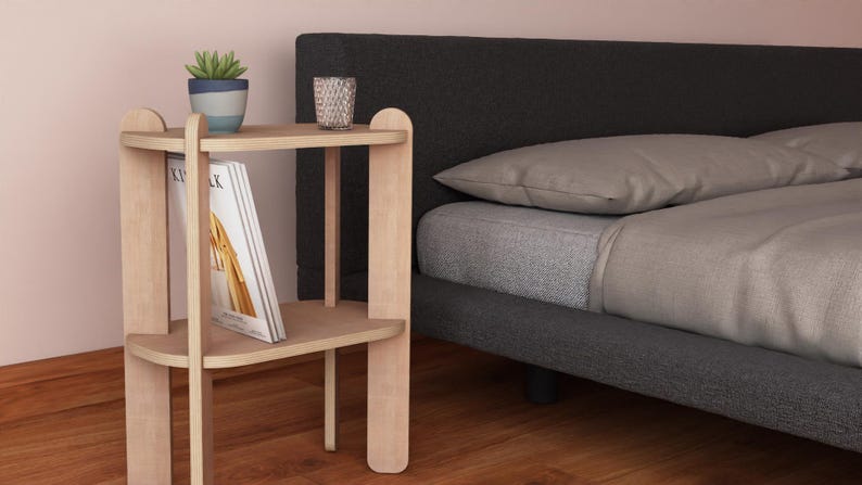 May include: A light wood bedside table with two shelves, holding a potted succulent, a glass, and a book. The table is next to a bed with gray bedding and a dark gray headboard.