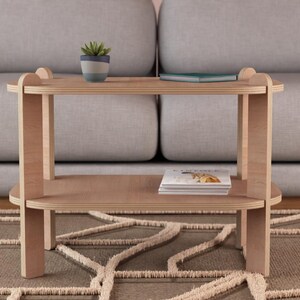 Puede incluir: Una mesa auxiliar de madera clara con dos estantes. Una peque&ntilde;a planta en maceta y un libro est&aacute;n en el estante superior, y una pila de revistas descansa en el estante inferior. La mesa est&aacute; frente a un sof&aacute; gris.