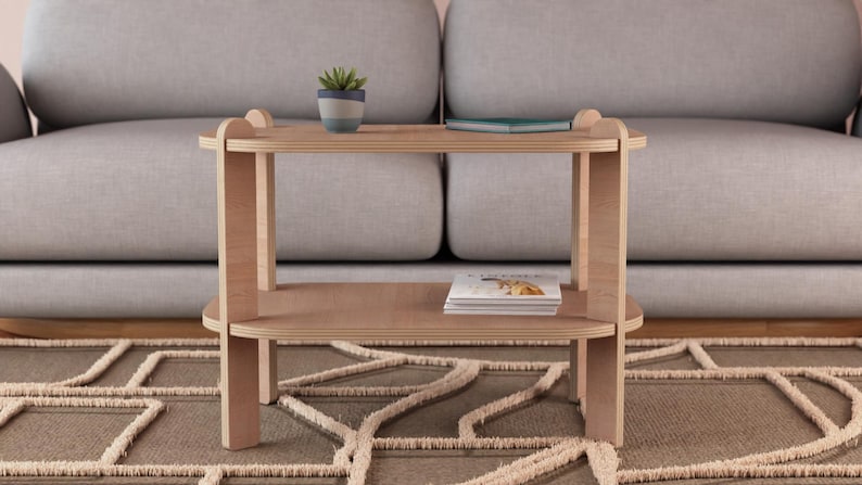 Puede incluir: Una mesa auxiliar de madera clara con dos estantes. Una peque&ntilde;a planta en maceta y un libro est&aacute;n en el estante superior, y una pila de revistas descansa en el estante inferior. La mesa est&aacute; frente a un sof&aacute; gris.