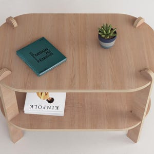 Puede incluir: Una mesa de centro de madera clara con un estante inferior. Un libro turquesa con el texto "DESIGN AU FEMININ" y una peque&ntilde;a planta en maceta est&aacute;n sobre la mesa. Una copia de "KINFOLK" est&aacute; en el estante inferior.