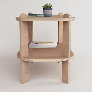 Puede incluir: Una mesa auxiliar de madera clara con dos estantes y esquinas redondeadas. Una peque&ntilde;a planta en maceta y un bol&iacute;grafo descansan sobre el estante superior. La mesa mide aproximadamente 61 cm de altura.