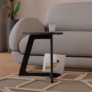 Peut inclure: Une table d'appoint noire avec un plateau arrondi et un design de pieds angulaires unique. La table est &agrave; c&ocirc;t&eacute; d'un canap&eacute; gris clair et a un livre et un verre sur le dessus.