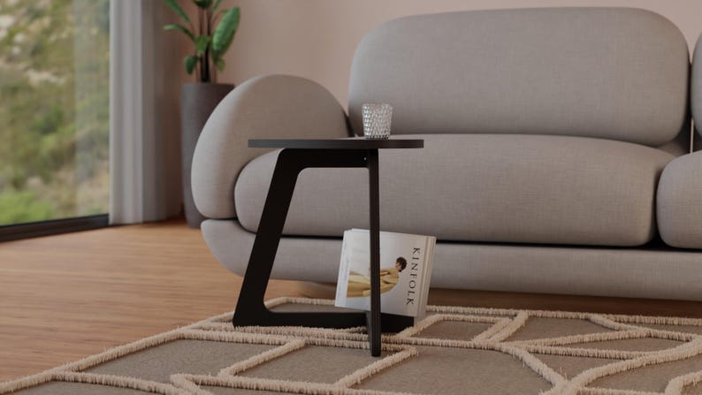 Peut inclure: Une table d'appoint noire avec un plateau arrondi et un design de pieds angulaires unique. La table est &agrave; c&ocirc;t&eacute; d'un canap&eacute; gris clair et a un livre et un verre sur le dessus.