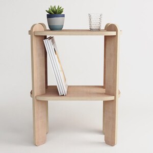 May include: A light-colored wooden side table with two shelves. A small potted plant and a clear glass sit on the top shelf. A book leans against the middle shelf. The table has a simple, modern design.