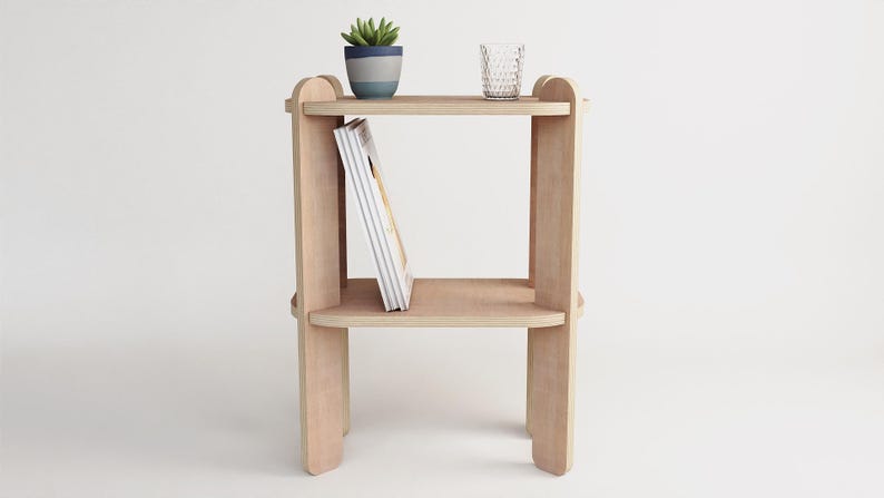 May include: A light-colored wooden side table with two shelves. A small potted plant and a clear glass sit on the top shelf. A book leans against the middle shelf. The table has a simple, modern design.
