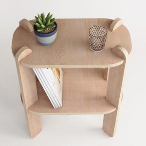 May include: A light-colored wooden side table with two shelves. A small potted plant and a decorative glass are on the top shelf. A stack of books is on the lower shelf. The table has a modern design.