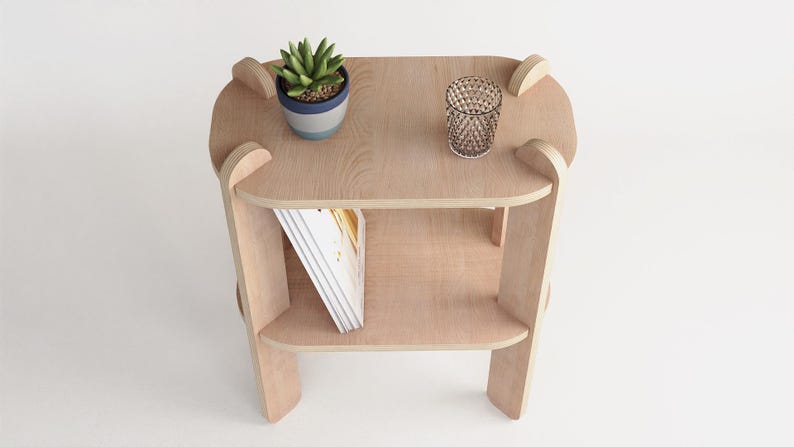 May include: A light-colored wooden side table with two shelves. A small potted plant and a decorative glass are on the top shelf. A stack of books is on the lower shelf. The table has a modern design.