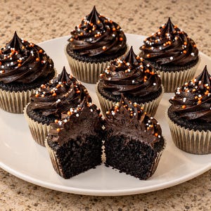 May include: Seven chocolate cupcakes with dark chocolate frosting and orange, white, and black sprinkles arranged on a white plate. One cupcake is sliced in half in the foreground, revealing the dark, porous texture of the chocolate cake.