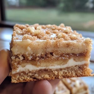 Puede incluir: Primer plano de una barra de postre cuadrada con múltiples capas. La base es una capa marrón claro y desmenuzable, cubierta con una capa de crema blanca, una capa de color caramelo y una parte superior cremosa y moteada. La parte superior está espolvoreada con migas.