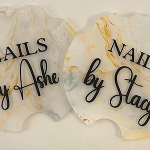 May include: Two white and gold marble-patterned coasters with black text. The first coaster says "Nails by Ashe" and the second coaster says "Nails by Stacy".