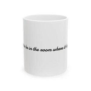 May include: A white ceramic mug featuring the black cursive text "to be in the room where it happens" printed horizontally across the center. The mug has a smooth, cylindrical body with a simple, clean design and a glossy finish throughout.