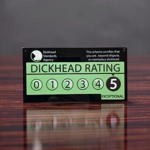 Op de afbeelding: Een rechthoekig zwart bord met groene accenten met de tekst Dickhead Standards Agency en This scheme certifies that you are, beyond dispute, an exemplary dickhead. Het bord toont een DICKHEAD RATING schaal van 0 tot 5.