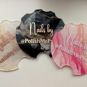 Op de afbeelding: Drie ronde acryl-onderzetters met een marmermotief. Elke onderzetter heeft de tekst "Nails by" en een social media-handvat. De onderzetters zijn in zwart, wit en goud, roze en goud, en wit en goud.