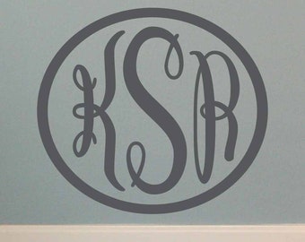 Könnte beinhalten: Ein grauer Monogramm-Aufkleber mit den Buchstaben "K S R" in einem Kreis. Der Aufkleber befindet sich an einer hellblauen Wand.