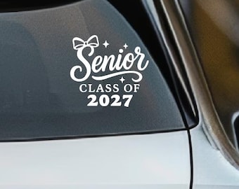 Könnte beinhalten: Ein weißer Vinyl-Aufkleber auf der Heckscheibe eines silbernen Fahrzeugs. Der Aufkleber zeigt den Text Senior CLASS OF 2027 in einer stilisierten Schreib- und Blockschrift, akzentuiert durch eine kleine Schleifengrafik und vierzackige Sterne.