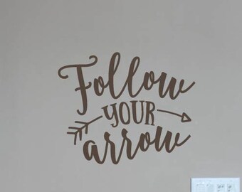 Könnte beinhalten: Brauner Wandaufkleber mit dem Schriftzug "Follow your arrow" in Schreibschrift, begleitet von einer Pfeilgrafik. Der Aufkleber befindet sich an einer beigen Wand, darunter Lichtschalter. Das Lively Lettering-Logo ist sichtbar.