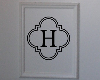 Könnte beinhalten: Ein gerahmtes Wandkunstwerk mit dem Buchstaben "H" in einem schwarzen, verzierten Design. Das Kunstwerk befindet sich vor einer weißen Wand mit rechteckigen Paneelen. Das Bild enthält einen Blick auf ein gemustertes Kissen.