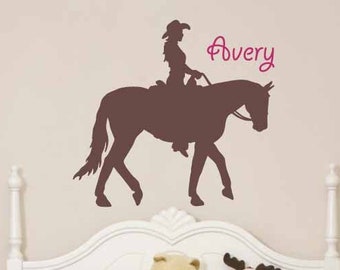 Könnte beinhalten: Eine braune Silhouette eines Cowboys auf einem Pferd ist an einer Wand über einem weißen Bett zu sehen. Der Name "Avery" ist in rosa Schrift geschrieben. Das Bett hat eine floral gemusterte Bettdecke und Kissen. Der Text "© Lively Lettering" steht unten.