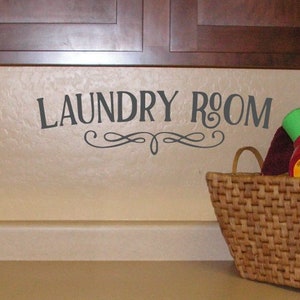 May include: A laundry room scene with a woven basket holding laundry supplies. The wall features the words "LAUNDRY ROOM" in a decorative font. The cabinets are dark brown, and the countertop is a light beige color.