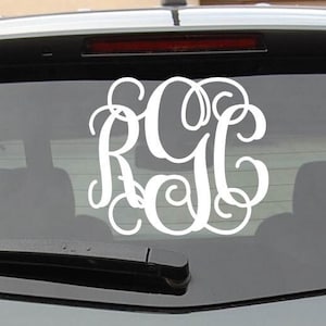 May include: A white monogram decal with the letters "RGC" in a cursive font, applied to the rear window of a gray car. The car has a gold Chevrolet emblem on the back.