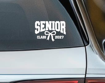 Könnte beinhalten: Ein weißer Vinyl-Aufkleber auf der Heckscheibe eines silbernen Fahrzeugs. Der Aufkleber zeigt den Text SENIOR in einer fetten Schriftart im College-Stil, wobei die Wörter CLASS und 2027 eine kleine, zentrierte Schleifengrafik flankieren.