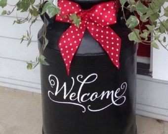 Könnte beinhalten: Eine schwarze Milchkanne aus Metall mit dem weißen Schriftzug "Welcome" in Schreibschrift. Ein rotes Band mit weißen Punkten ist um den Hals gebunden. Eine grüne Efeupflanze steht darauf. Der Text "DIY- Milkcan makeover An Easy front porch project!" ist sichtbar.