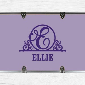May include: A rectangular lavender storage trunk featuring a dark purple monogram of the letter E enclosed in a circular decorative frame. The name ELLIE is printed in dark purple block letters below the monogram. The trunk has silver metal edges.