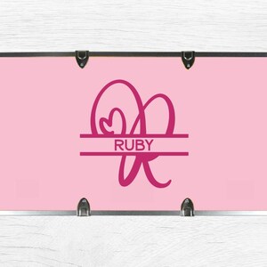 May include: A rectangular light pink storage trunk featuring a metallic silver frame with corner reinforcements. The center displays a dark pink monogram of the letter R with a heart shape and the name RUBY printed in a horizontal line.