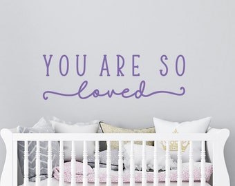 Könnte beinhalten: Lilafarbener Vinyl-Wandaufkleber mit dem Text "You are so loved" in einem weißen Babybett mit rosa und grauem Bettzeug.