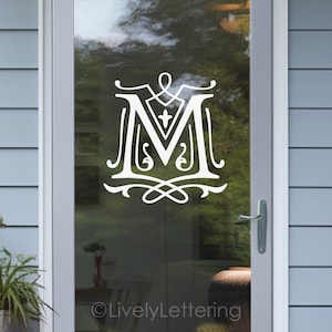 May include: White vinyl decal of a monogram letter M with decorative flourishes. The decal is applied to a glass door. The text "©LivelyLettering" is visible at the bottom of the image.