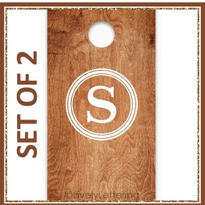 May include: Set of 2 wooden cornhole boards with a brown wood grain finish. Each board features a white circular design with the letter "S" and a hole at the top. The words "SET OF 2" are printed vertically on the left side.
