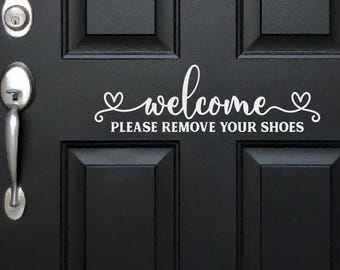 Könnte beinhalten: Ein weißer Vinyl-Aufkleber auf einer schwarzen Tür, der das Wort welcome in Schreibschrift zeigt, flankiert von zwei Herzumrissen, mit dem Text PLEASE REMOVE YOUR SHOES in einer klaren, serifenlosen Schrift direkt unter dem Wort welcome.