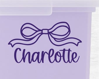 Könnte beinhalten: Eine hellviolette Kunststoff-Aufbewahrungsbox mit einem durchscheinenden Deckel. Auf der Vorderseite befindet sich eine dunkelviolette Grafik einer dekorativen Schleife über dem Namen Charlotte in Schreibschrift. Der Text LivelyLettering ist sichtbar.