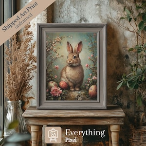 May include: A framed art print featuring a brown rabbit sitting in a field of pink flowers with Easter eggs. The background is a soft blue with a light blue sky. The print is in a gray frame.