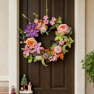 以下が含まれることがあります： 様々な色の花と葉が特徴の手作りのかぎ針編みのリース。紫、ピンク、オレンジ、黄色の花と、緑の葉と茎が含まれています。濃い茶色のドアに掛けられています。