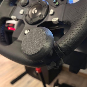 Könnte beinhalten: Nahaufnahme eines schwarzen Rennlenkrads mit strukturiertem Griff und einem schwarzen Knopf auf der rechten Seite.