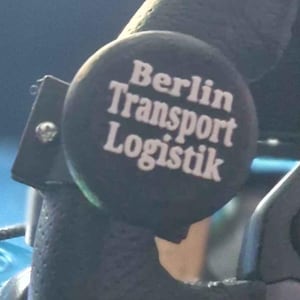 Könnte beinhalten: Nahaufnahme eines schwarzen Rennlenkrads mit strukturierter Grifffläche. Das Lenkrad hat blaue Knöpfe und ein PlayStation-Logo. Ein schwarzer Knopf mit dem Text "Berlin Transport Logistik" ist am Lenkrad befestigt.
