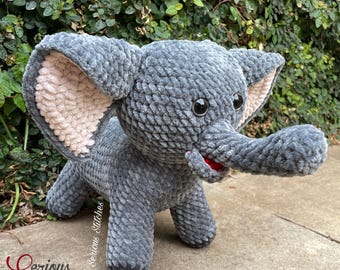 Könnte beinhalten: Ein graues gehäkeltes Elefanten-Plüschtier mit großen Ohren, einem Rüssel und kleinen schwarzen Augen. Die Innenseite der Ohren ist hellbeige und der Mund ist rot. Der Elefant steht auf einer Betonoberfläche vor grünem Laub.