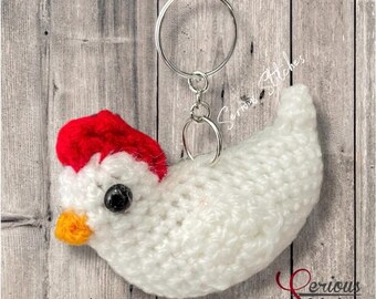 May include: A handmade white crochet chicken keychain with a red hat and a yellow beak. The chicken has a black bead eye and a silver key ring. The keychain is a unique, handcrafted accessory.