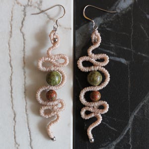 May include: A pair of long, pink, beaded earrings with green and orange stones. The earrings are made of a woven cord and have a silver hook.