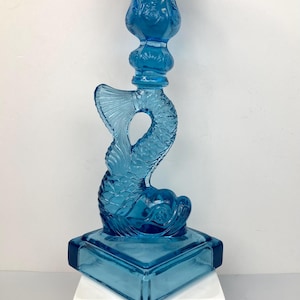 May include: A blue glass candlestick holder with a fish design. The candlestick features a fish tail and body, and a square base. The top of the candlestick has a floral design. The candlestick is made of transparent blue glass.