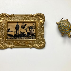May include: A vintage gold-toned metal picture frame with an ornate design, holding a black and white photograph of people near a boat. A small, decorative turtle figurine with blue and gold accents is also present.