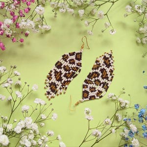 May include: Two beaded leopard print earrings in brown, white, and tan, with gold-coloured hooks. The earrings are teardrop-shaped. They are displayed on a light green surface, surrounded by white, pink, and blue flowers.