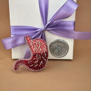 Könnte beinhalten: Handgefertigte, perlenbesetzte Brosche in Form eines Magens, in Rot- und Rosatönen. Sie liegt neben einer weißen Geschenkbox mit lila Schleife und einer silbernen Münze.