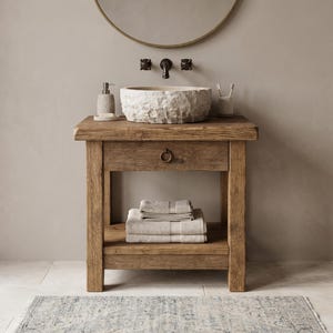 May include: A wooden bathroom vanity with a stone basin sink, a circular mirror, and a cabinet. The cabinet has a drawer and a lower shelf with folded towels. A soap dispenser and toothbrush holder are on the countertop. The rug has a blue and white pattern.