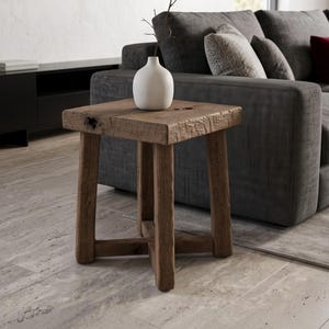 May include: A square wooden side table with a rough, weathered texture and four sturdy legs connected by a cross-brace base. A small, smooth white ceramic vase containing a single thin, bare branch sits on the flat top surface of the table.